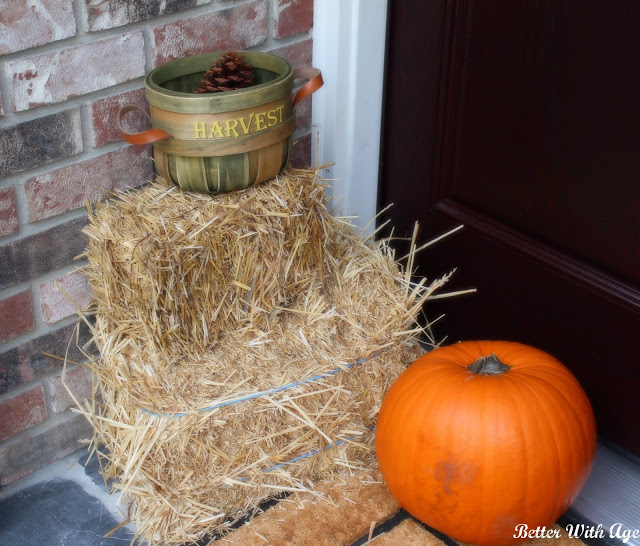 A bale of hay, a harvest bucket with a pine cone in it and an orange pumpkin on porch.