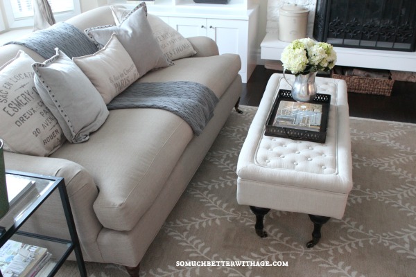 A neutral couch with a throw blanket and pillows plus a white ottoman with flowers in a vase.