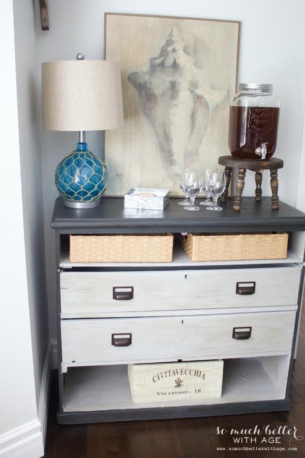 A small console table with wine glasses and a beachy lamp on top.