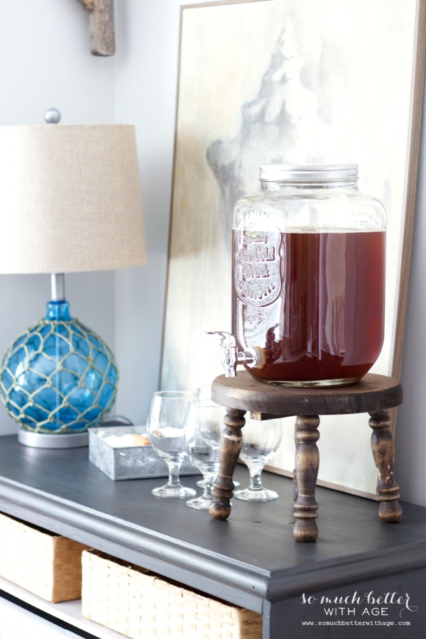 A clear glass beverage holder with a spout for pouring and glasses beside it.