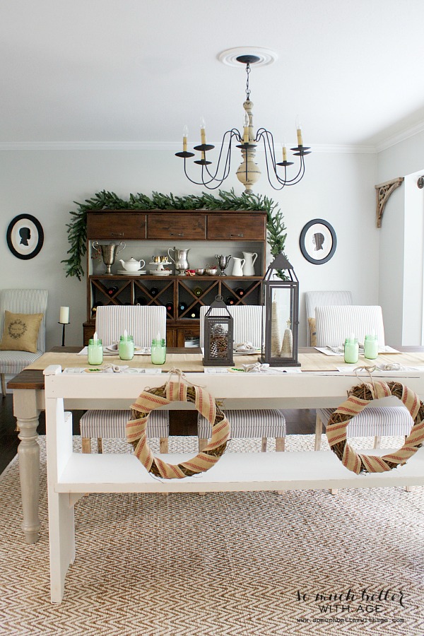 Christmas wreaths hanging on the back of white bench at dining table and open shelving behind table.