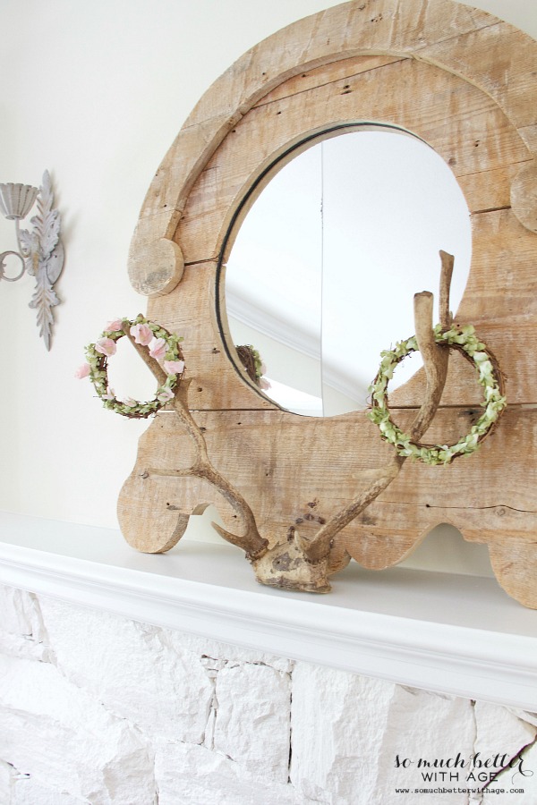 A white mantel with a wooden mirror and two floral crown on the mantel.