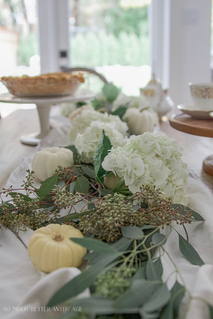 White flowers, and seeded branches with white pumpkins on a white table runner.