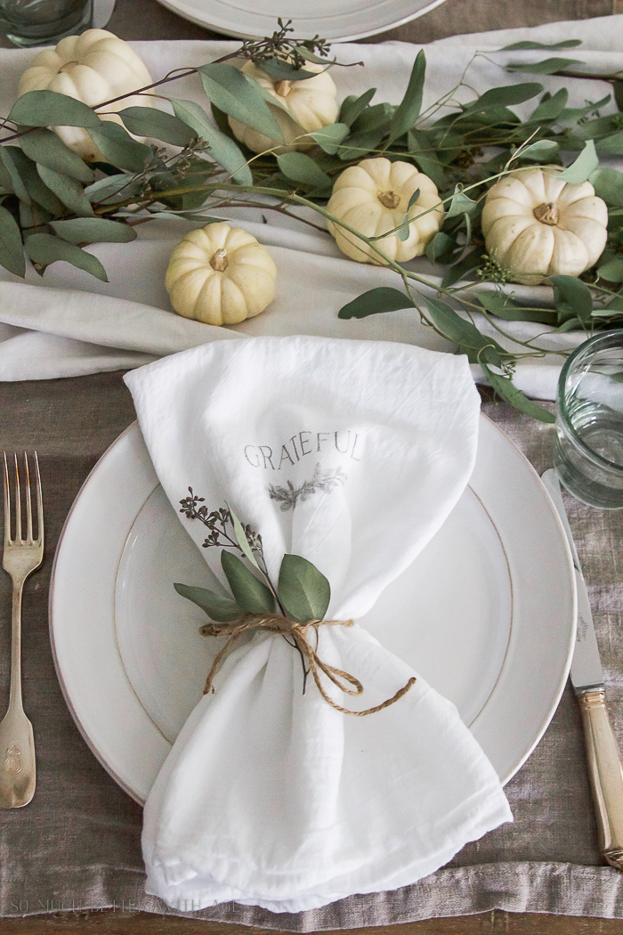 The table set for Thanksgiving with a napkin on a white plate and white pumpkins and leaves on the table.