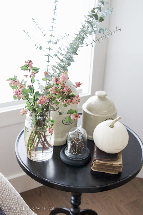 A small black round table with a glass jar filled with greenery and pink berries, a vintage book, a white pumpkin.