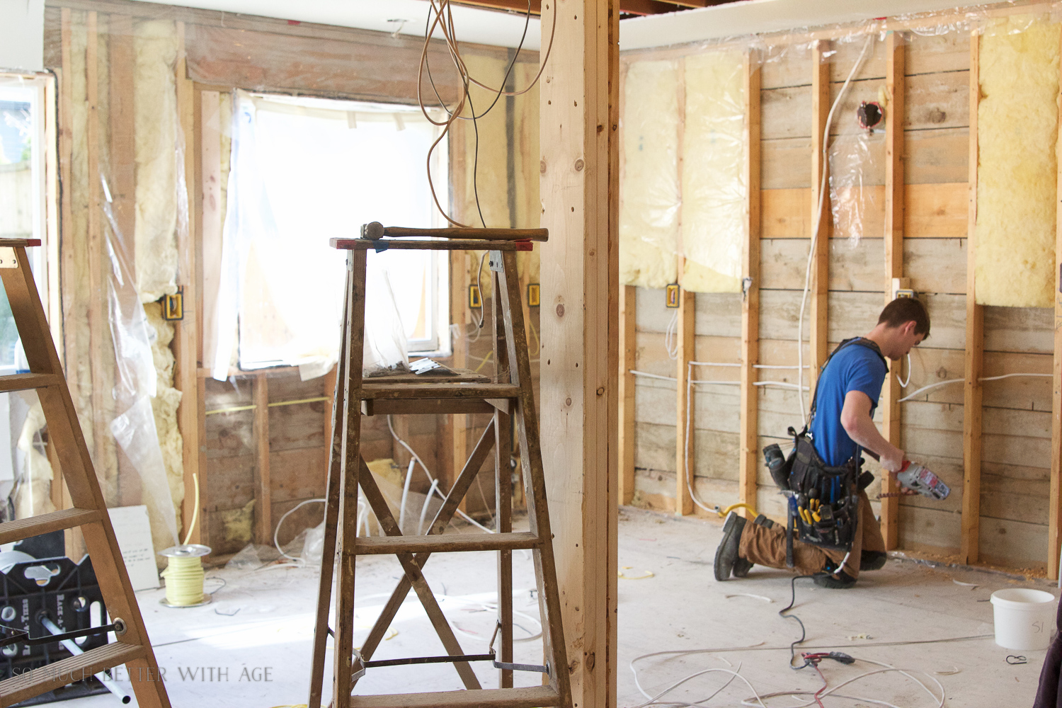 A construction worker is on his knees drilling and a ladder is in the center of the room.