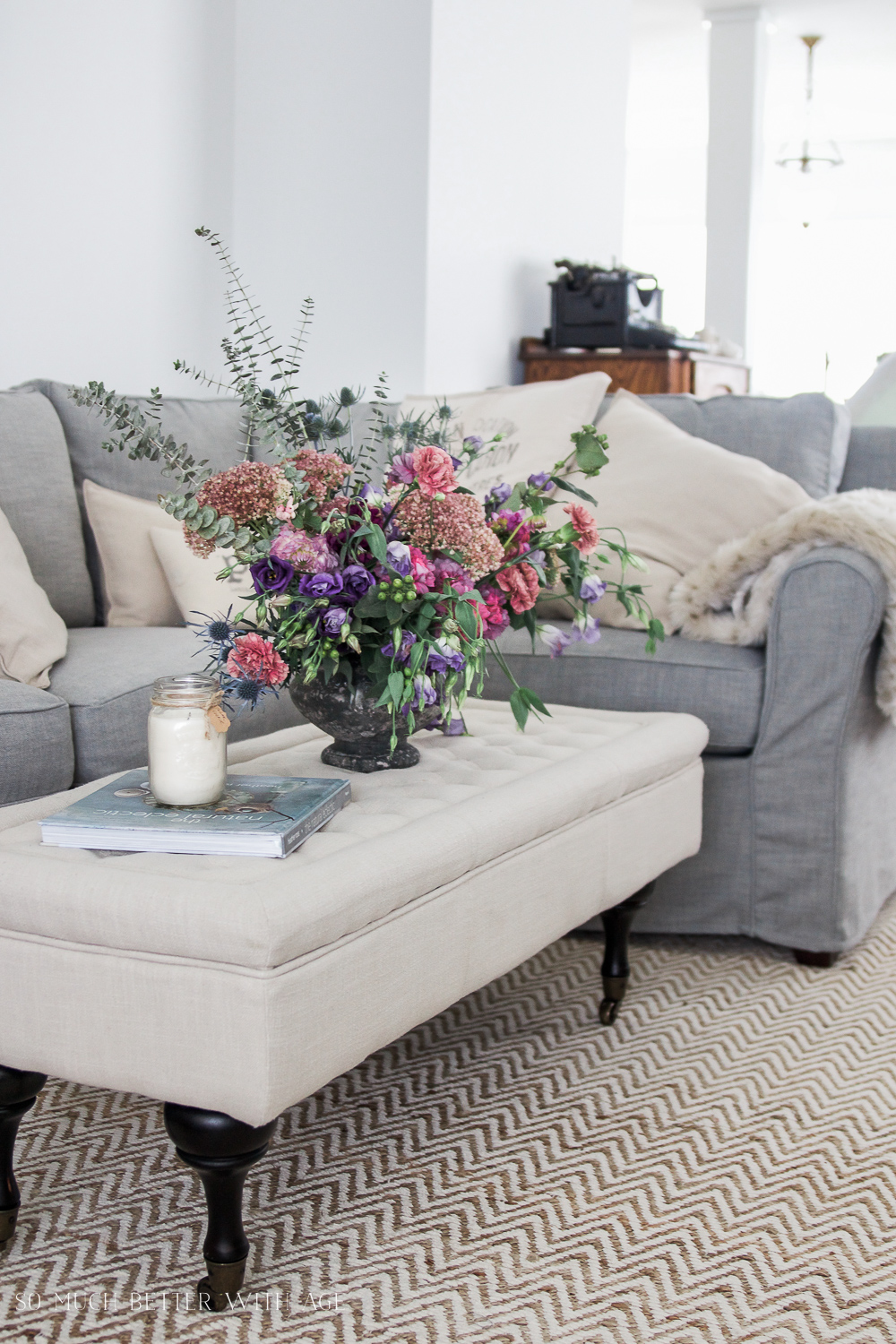 A tufted neutral ottoman with a large display of flowers on it, and gray couch behind it.