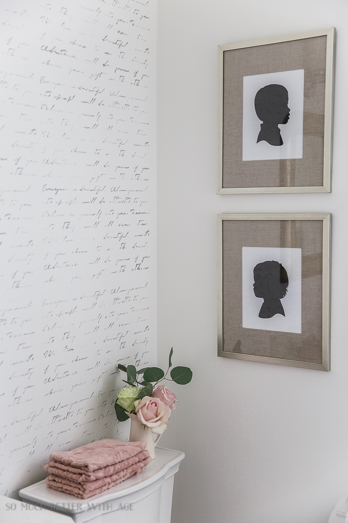 There are pink and white flowers in a white vase on the back of the toilet.