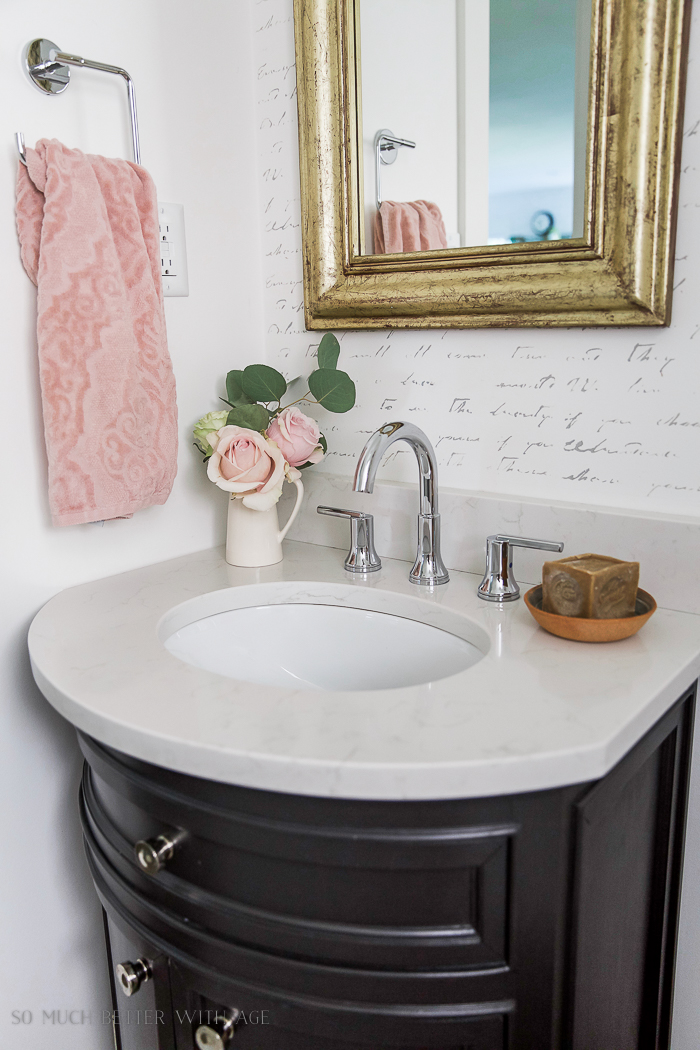 Gold mirror, little sink, pink towel and pink flowers in small bathroom.