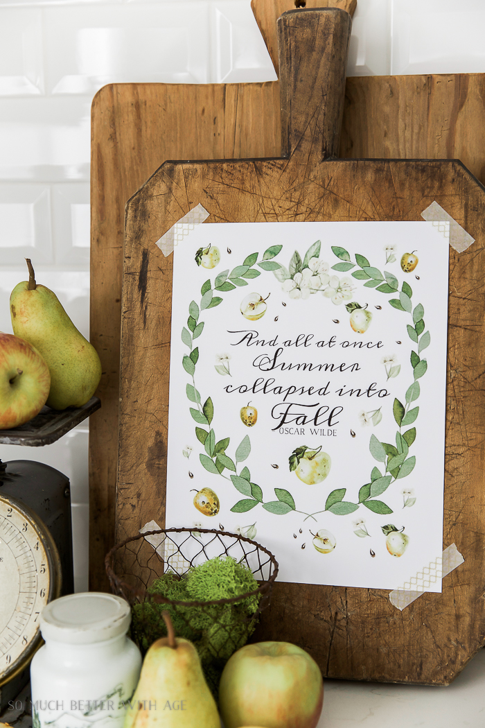 Apples and eucalyptus free fall printable/ pears, French bread board and a wire container filled with moss in front of it in white kitchen.