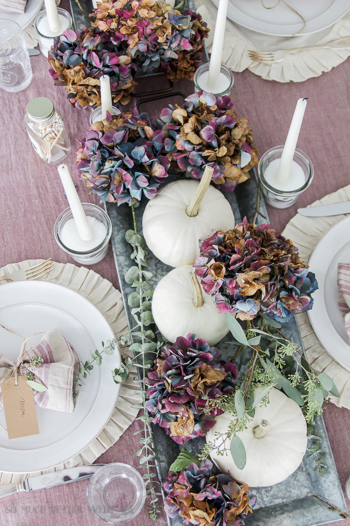 Dried blue and purple hydrangeas on the table with white candles and white plates.