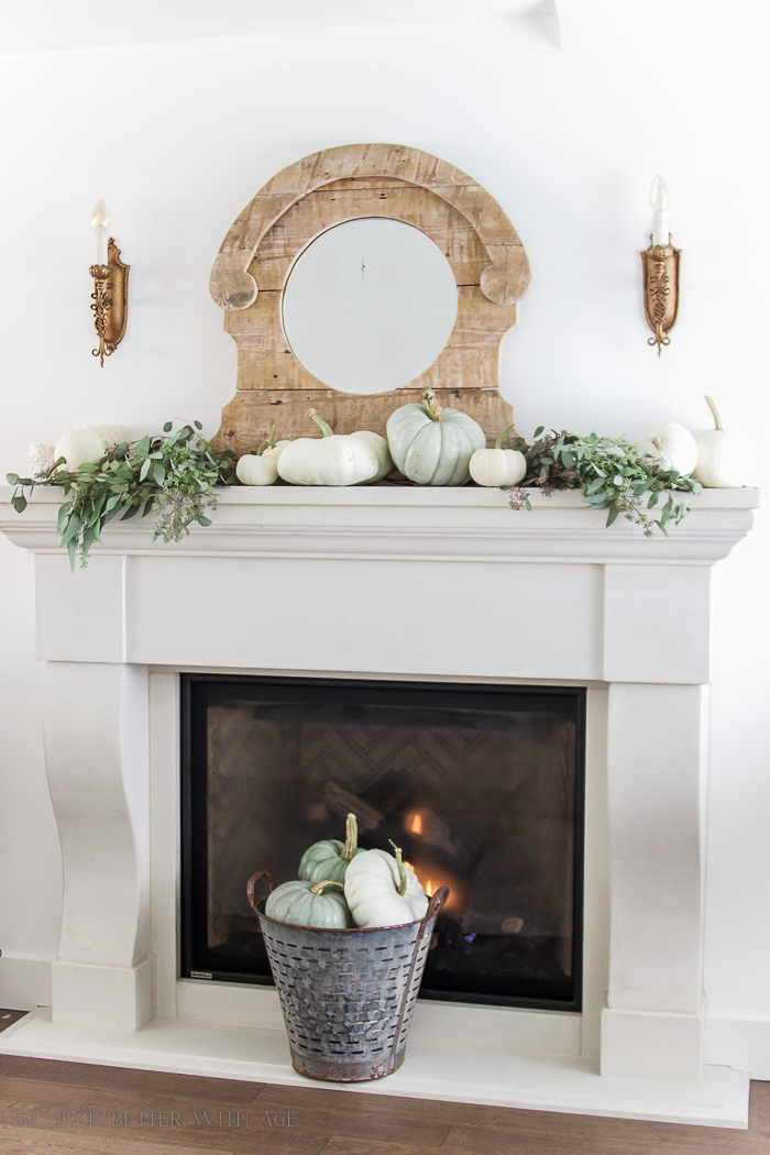 White neutral fireplace with mirror above it and pumpkins on mantel.