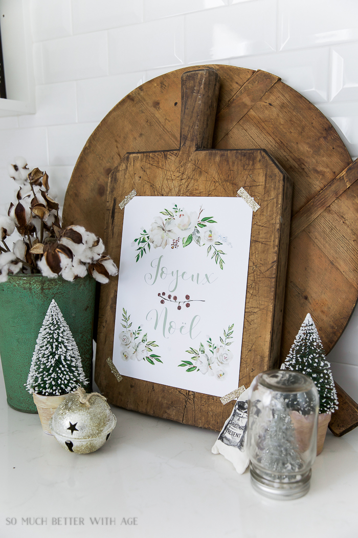 Joyeux Noel - Free Christmas Printable/Merry Christmas on the bread board in the kitchen surrounded by mini Christmas trees o the counter.
