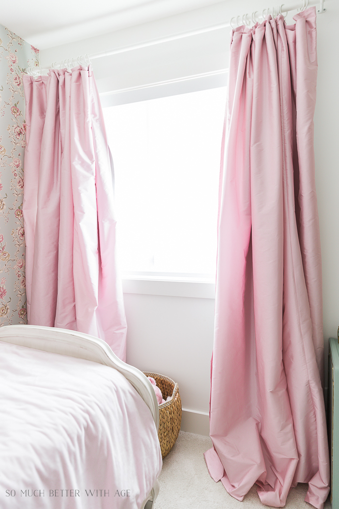The soft pink curtains in the bedroom with a basket underneath the window in the bedroom.