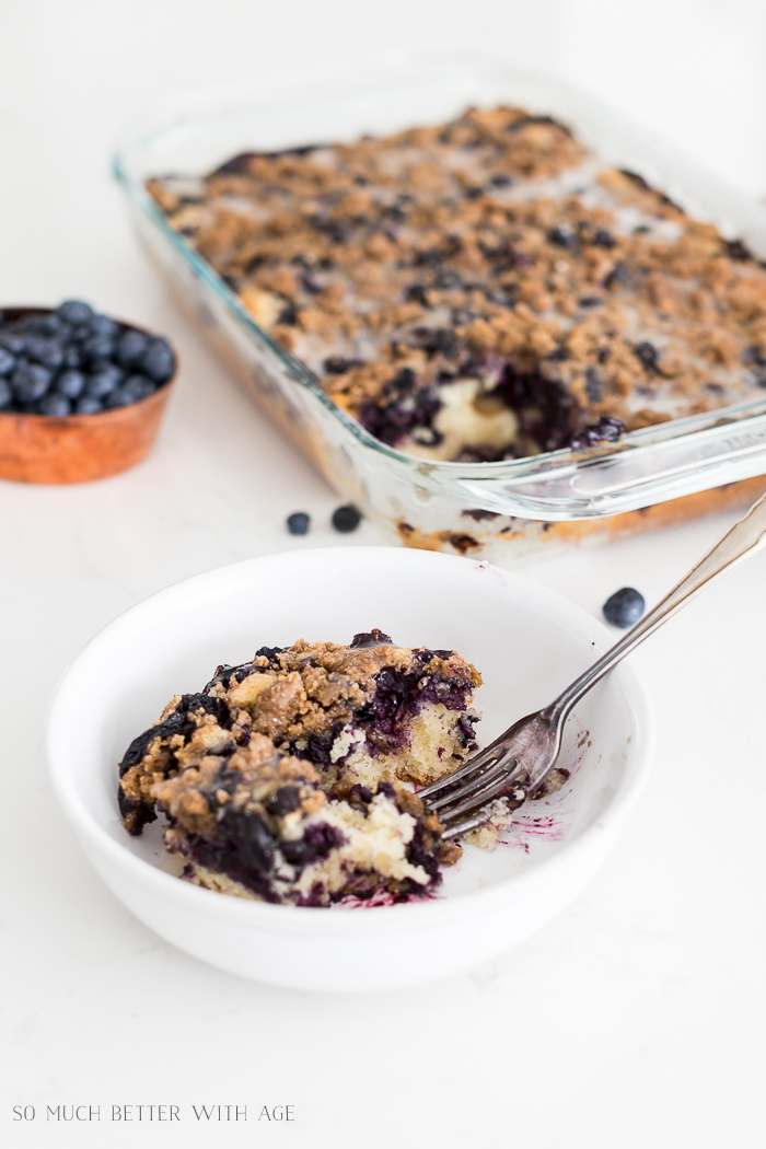 The baked blueberry streusel crumb cake in a clear dish with a piece in a bowl and a fork in it.