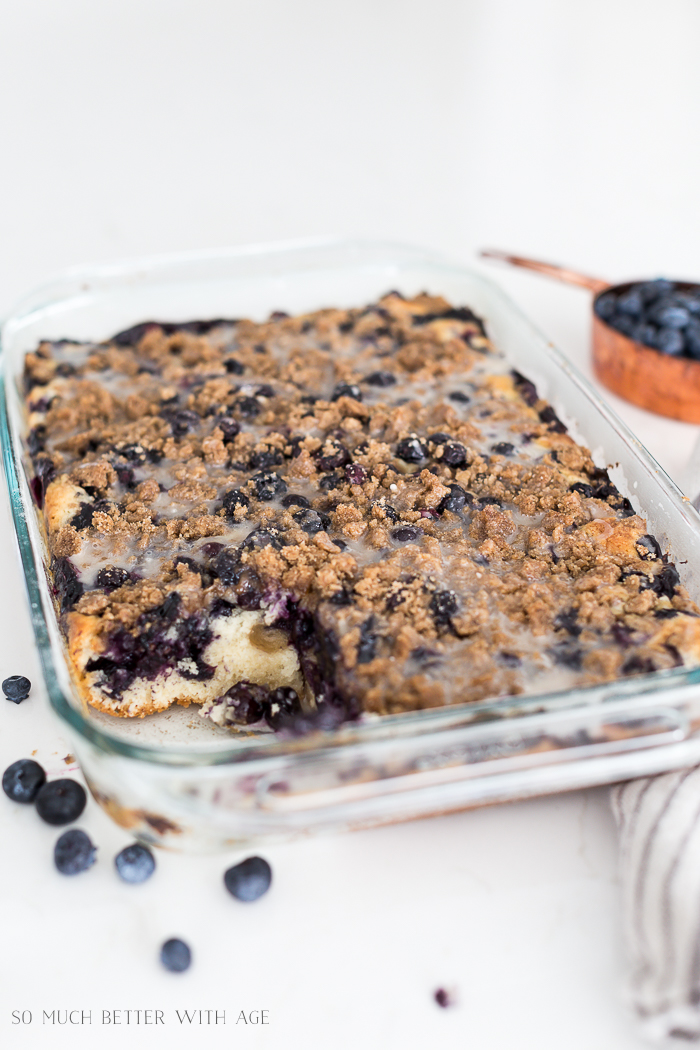 The baked blueberry crumb cake with a piece missing on a counter with blueberries around it.