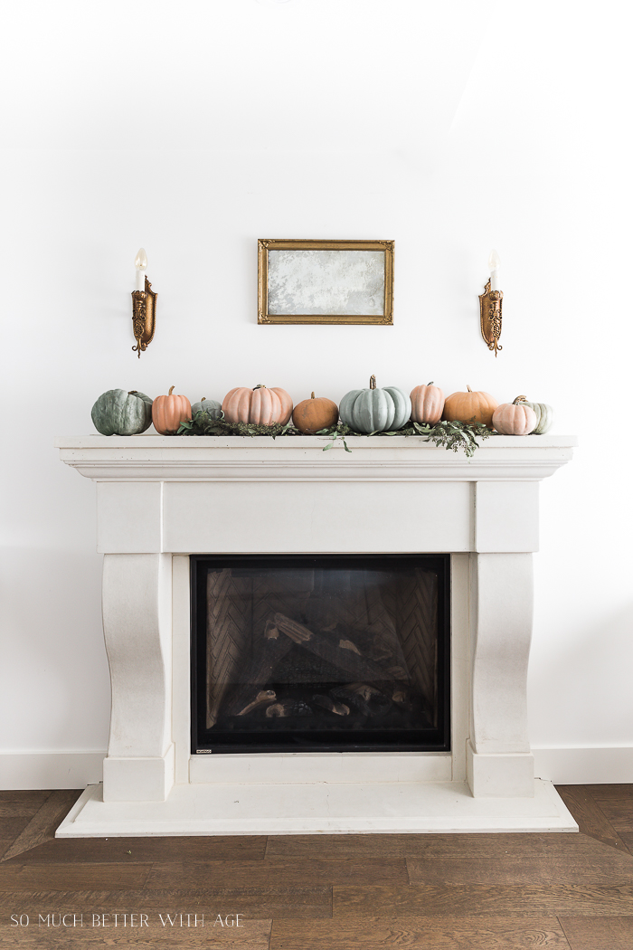 White fireplace mantel with soft colored pumpkins flanked by gold wall sconces.