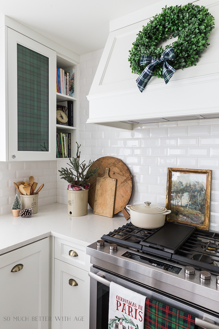 Pinecones on the counter, bread board and wooden cutting board, a white pot on the stove and plaid cloth in the cupboard where there is glass.