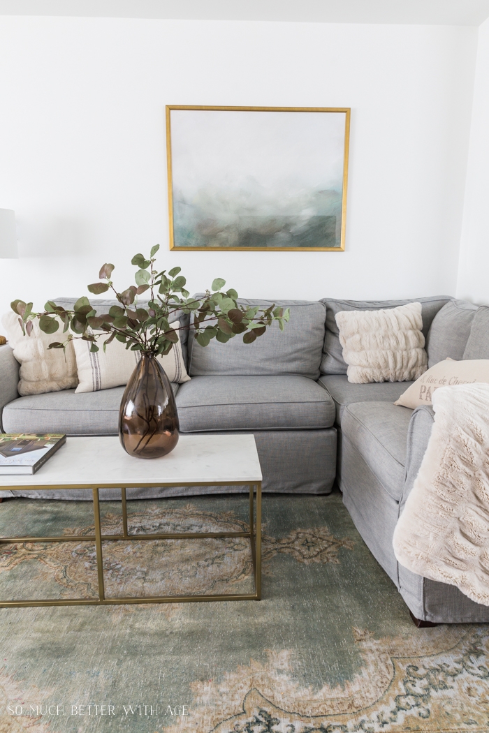 Grey couch, green rug, green and gold artwork and brown vase with greenery on marble and gold coffee table. 