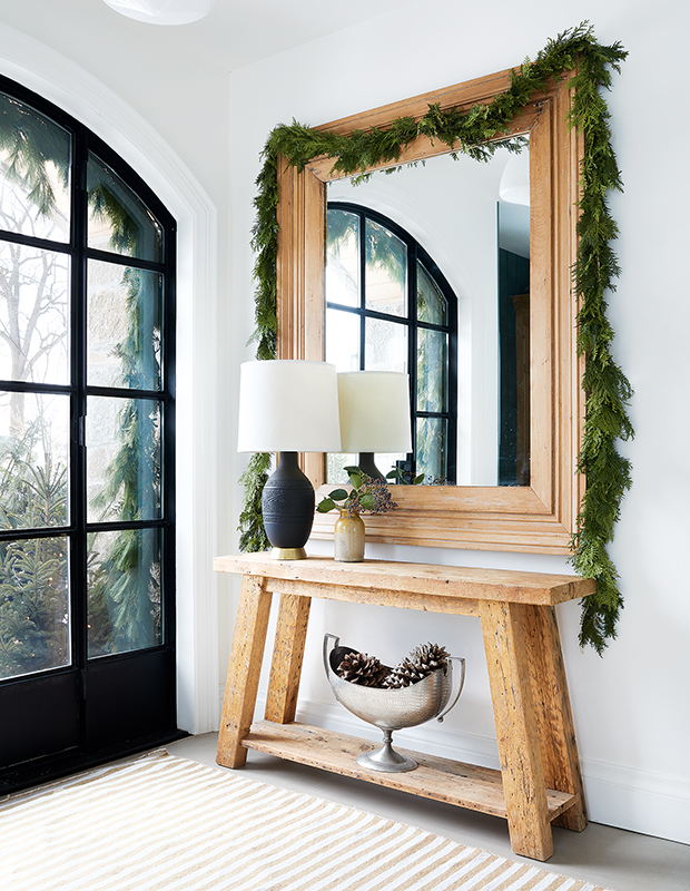Green garland hung over a large mirror in foyer with black glass door. 