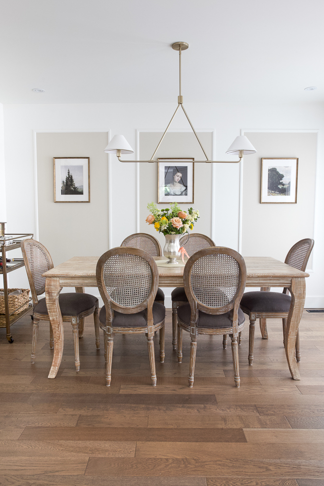 Gold modern light fixture above French dining table and chair set with floral arrangement on top of table.