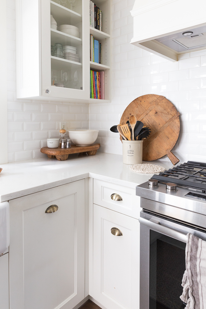 White kitchen with white subway tile walls, glass front cabinet and wooden round cutting board leaning on tiled wall.