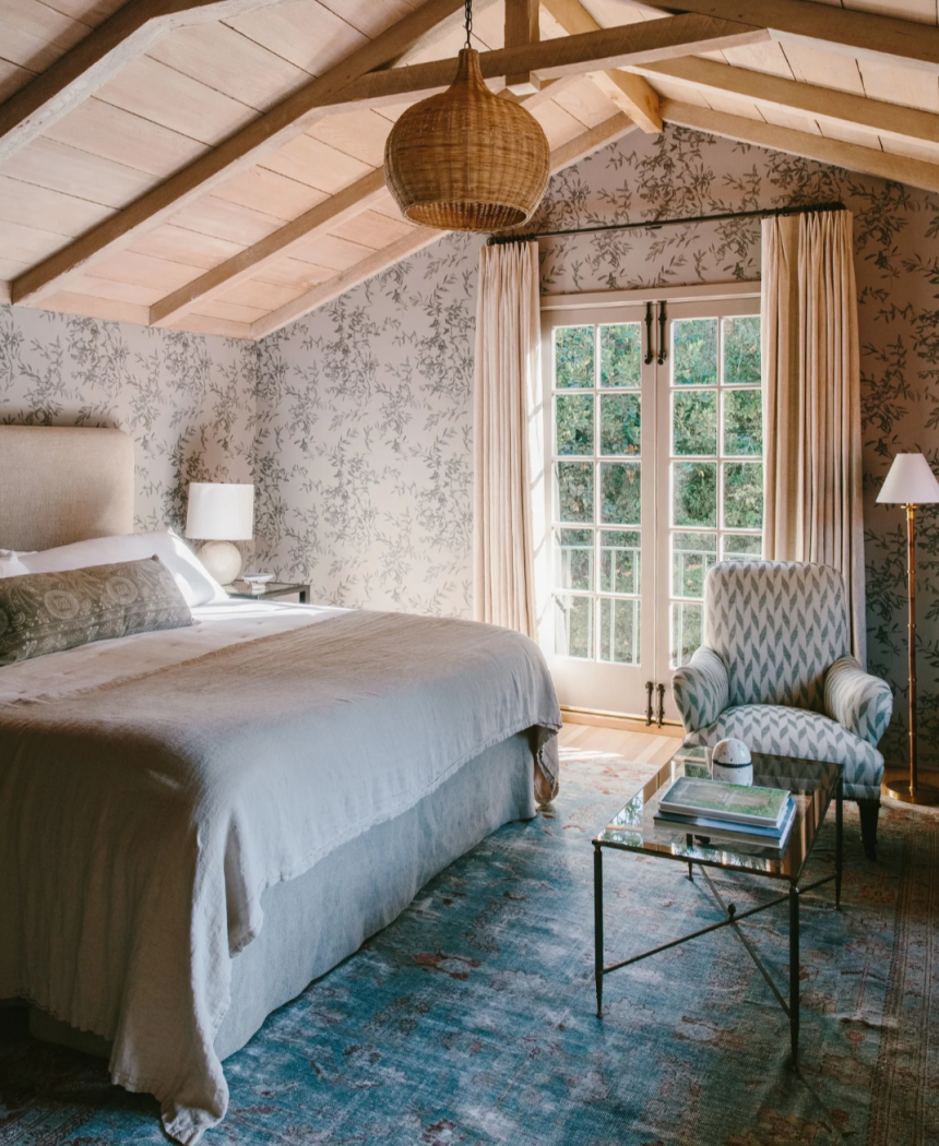 Bedroom with linen bedding, floral wallpaper and wooden vaulted ceiling.