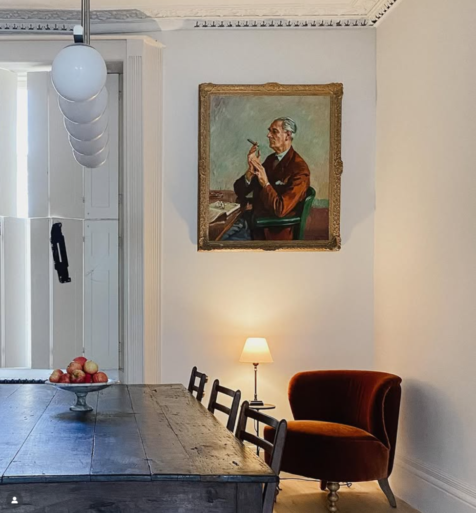 Oil painting on wall with vintage dining table and chairs and velvet armchair in corner. 