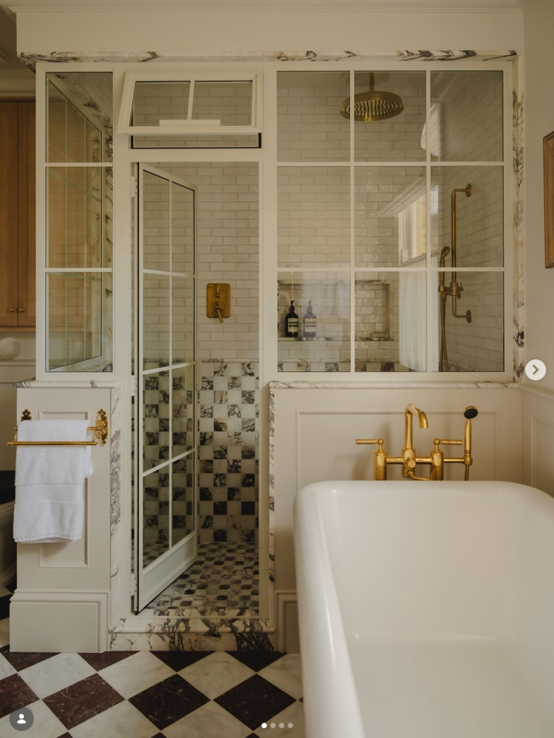 Marble checkerboard tiles in bathroom with glass and metal cream shower door and windows and freestanding tub with gold faucet.